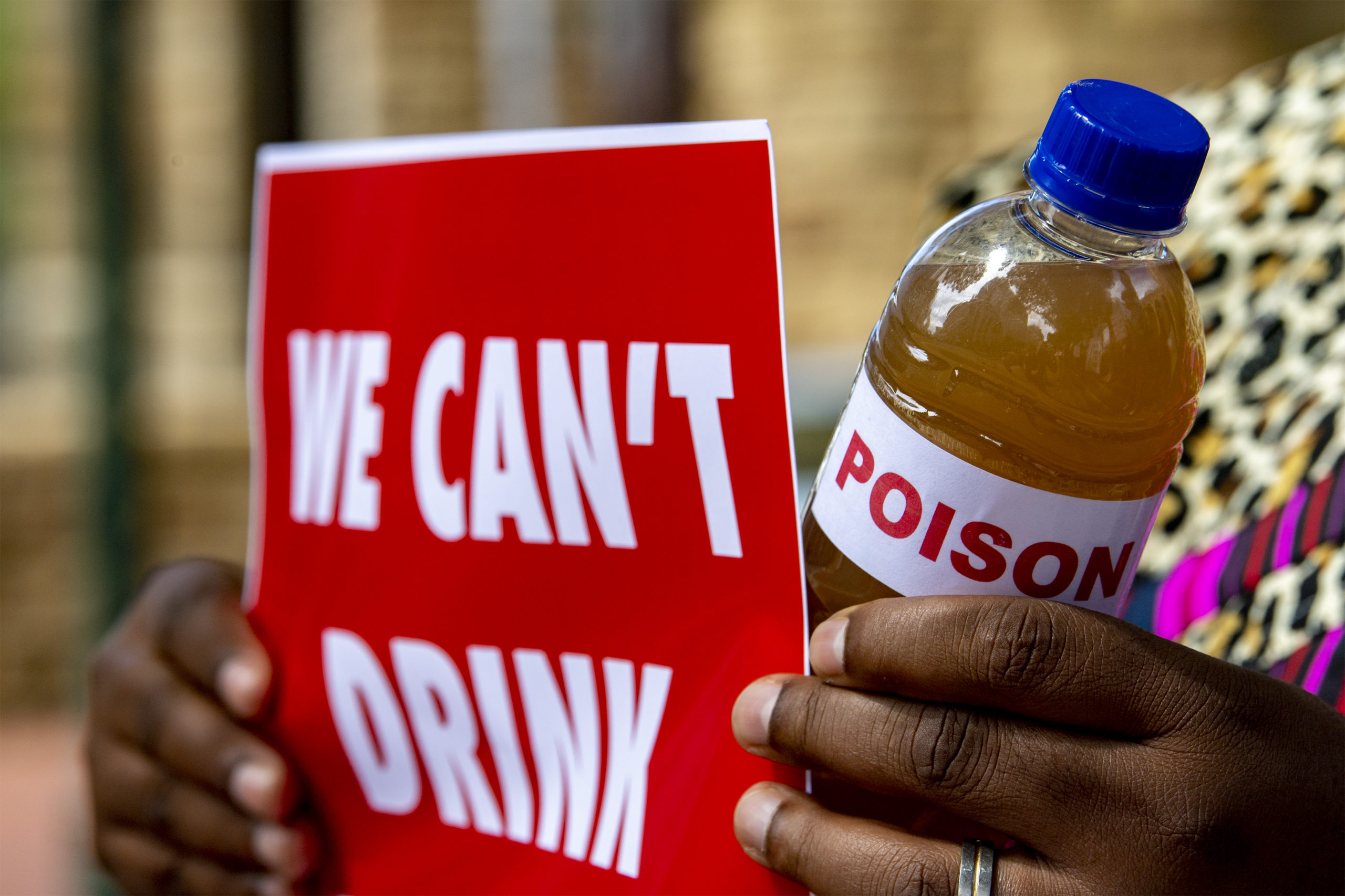 A picture of a sign that reads "WE CAN'T DRINK" and a bottle of dirty water with a "poison" label on it held by a person at a silent protest against the drilling in the Kavango Basin on the steps of St. George's Cathedral.